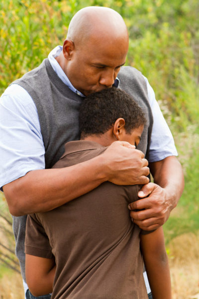 African-American father hugging his teenage son in an outdoor setting